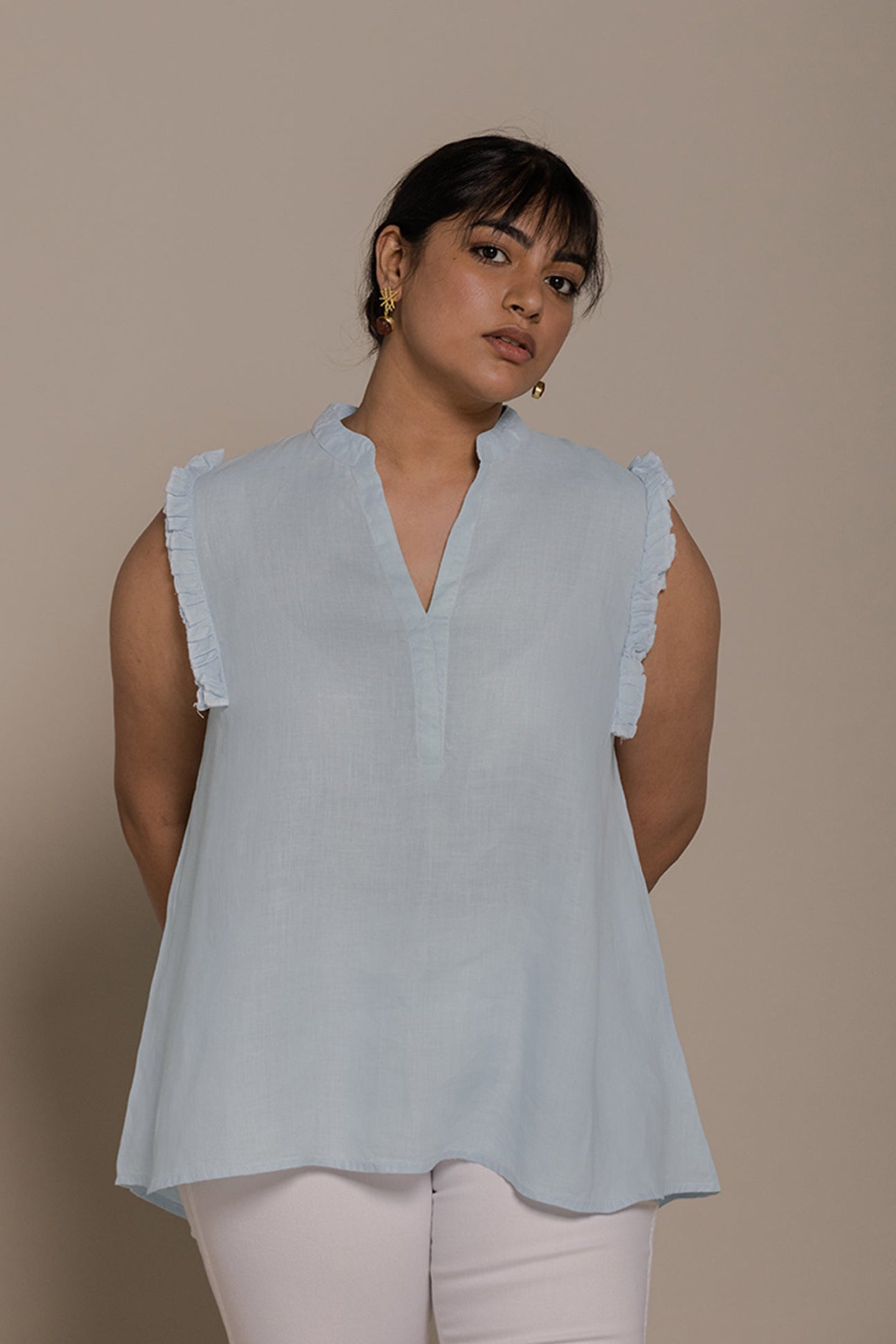 A female model wearing a blue, ruffle sleeveless top with a mandarin v-neck collar, paired with white pants.